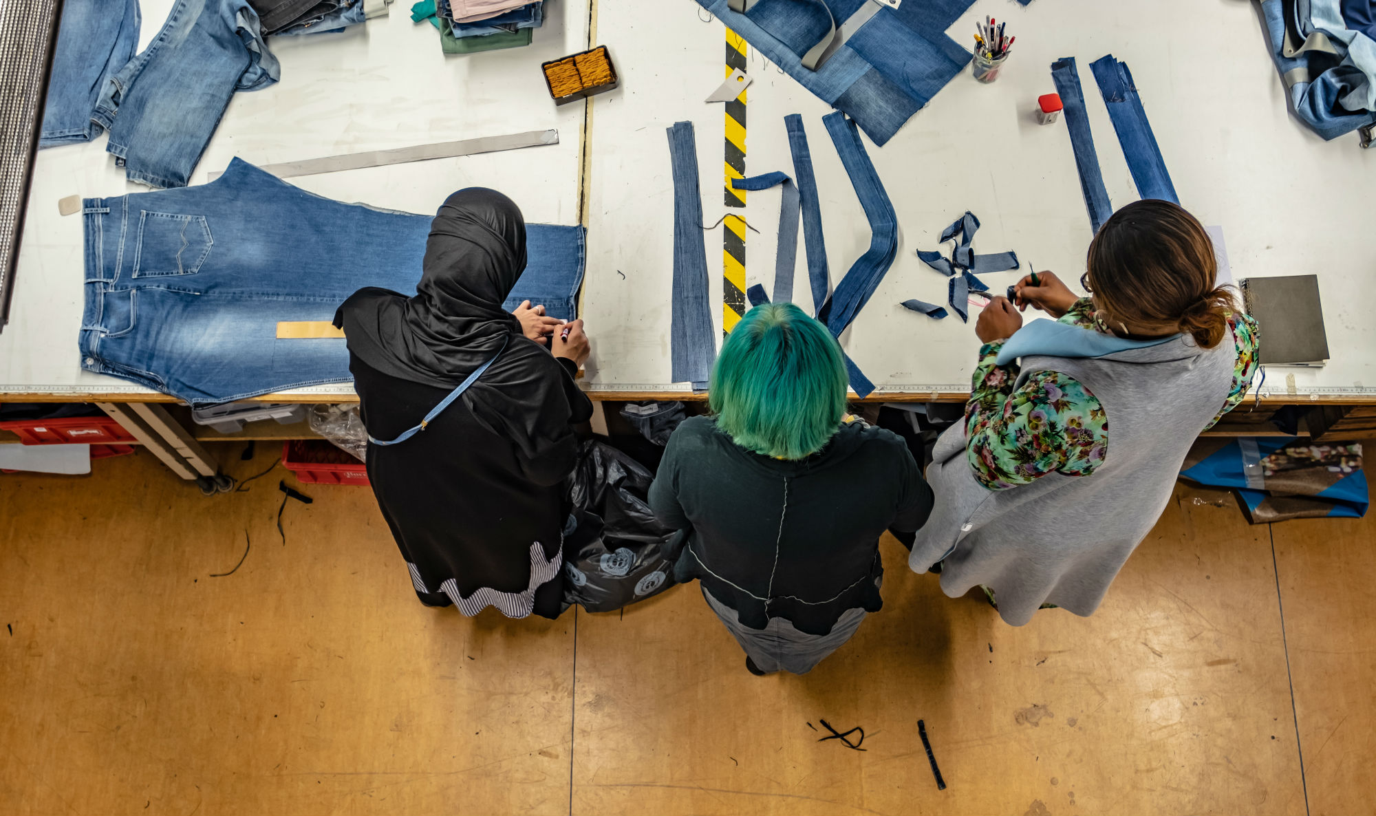 Vielfältiges Produktionsteam der Bridge&Tunnel GmbH Hamburg bei der nachhaltigen Upcycling-Arbeit mit Jeansstoffen in der Textilmanufaktur. Drei Mitarbeiterinnen mit unterschiedlichen kulturellen Hintergründen schneiden und verarbeiten Denim-Materialien zu neuen Designerstücken - ein anschauliches Beispiel für soziale Integration und nachhaltige Modewirtschaft.