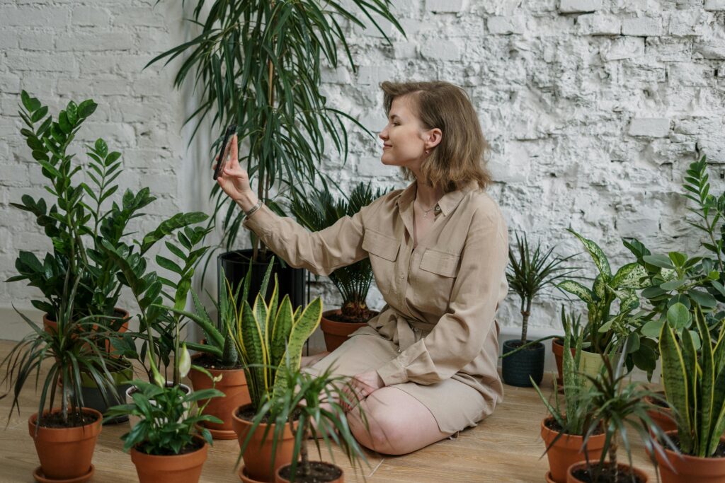 Junge Frau in beigefarbener Kleidung umgeben von einer Sammlung verschiedener Zimmerpflanzen vor weißer Backsteinwand – Indoor-Gardening als Teil der urbanen Gartenkultur