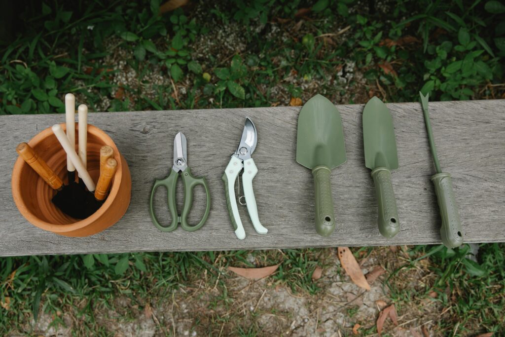 Gartengeräte für Urban Gardening auf einer Holzbank: Gartenscheren, Handschaufeln und Pflanzstäbe neben einem Tontopf mit Markierungsstäbchen – Werkzeuge für nachhaltige Stadtgärtnerei