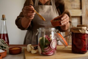 andwerkliche Fermentation: Person in Schürze gibt Gewürze zu geschnittenem Radieschen und Dill in Einmachglas, umgeben von fermentierten Produkten in nachhaltigen Gläsern